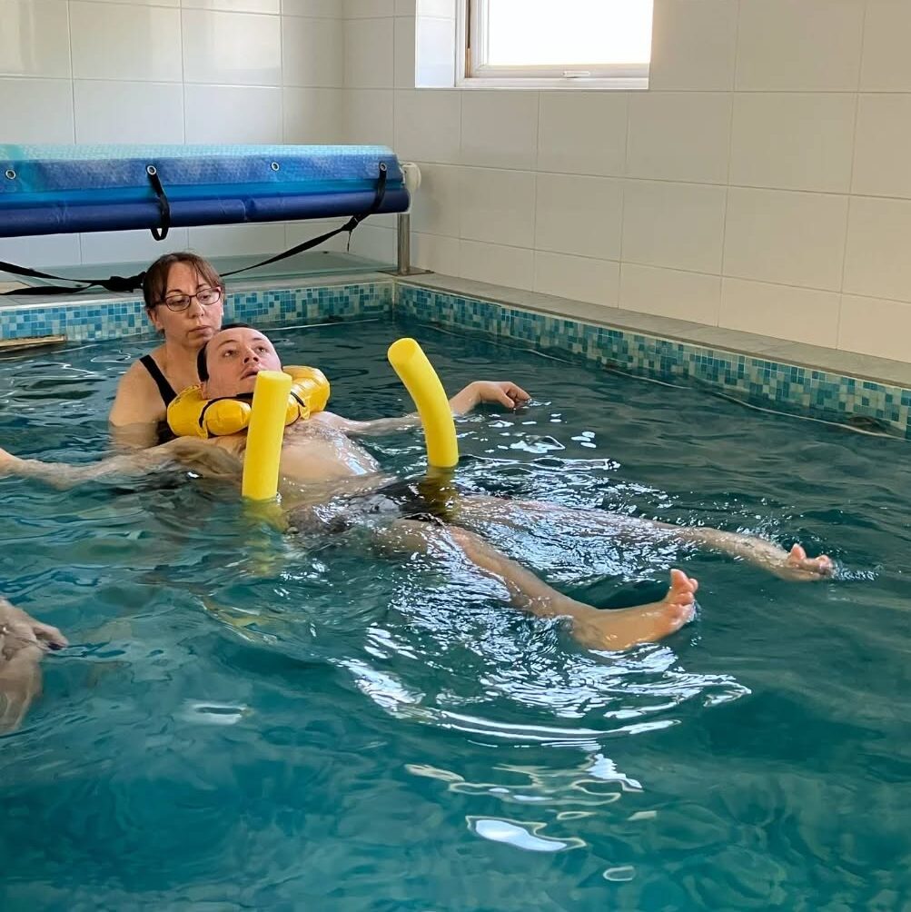 James is on his back in the pool, supported by a person, and with yellow tube floats.