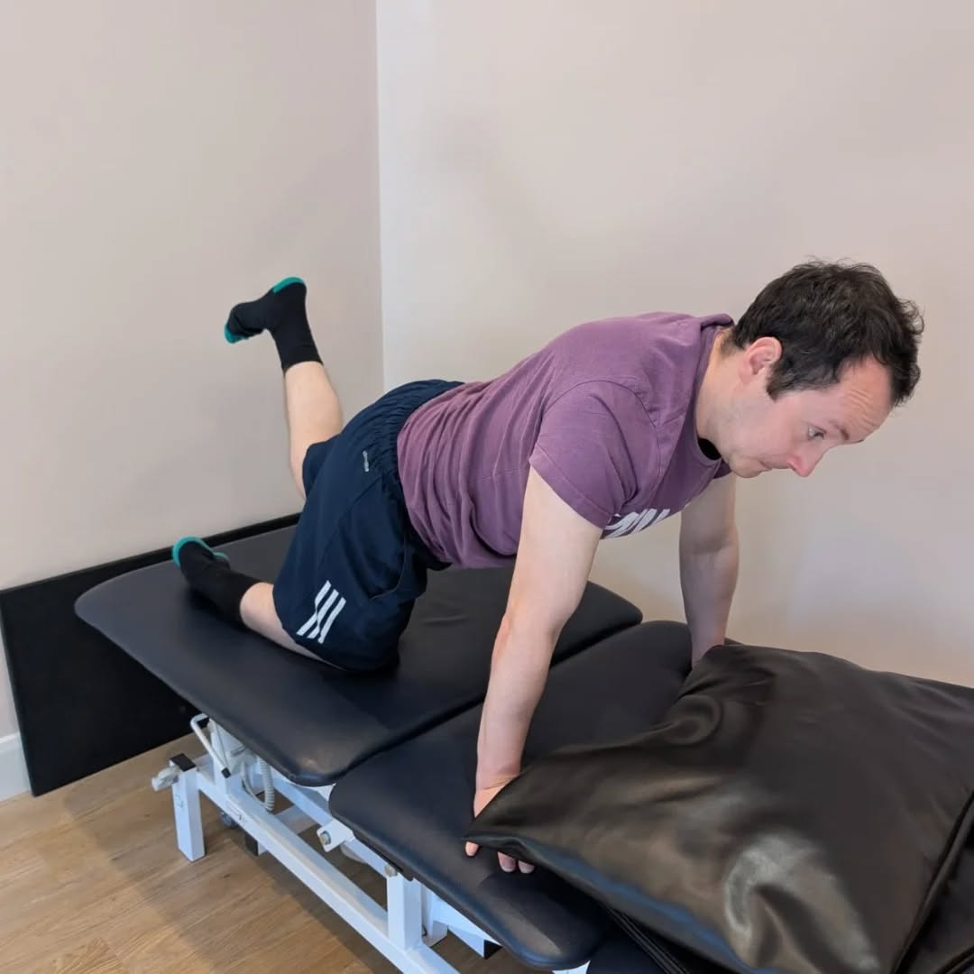James is kneeling on one knee on a physio bed. More rehab.